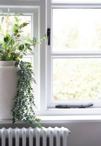 From The Anvil's Aged Bronze Hinton window fastener & window stay on a white window frame, with a large ceramic pot with green plants on the windowsill and a painted white radiator below.