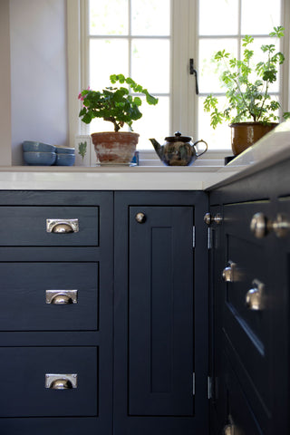 From The Anvil's Polished Nickel Art Deco drawer pulls on a navy kitchen cabinet with a white countertop covered in potted plants.