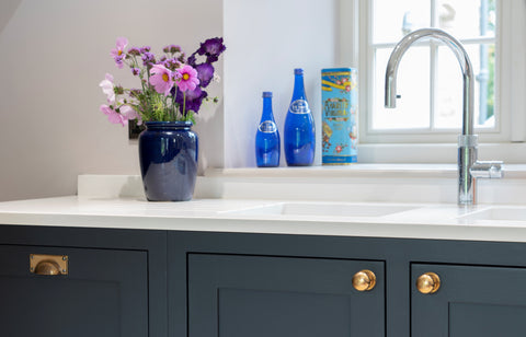 From The Anvil's Aged Brass Hammered cabinet knob & drawer pull on navy blue cabinets with a white marble countertop, steel sink, and blue pots with pink flowers.