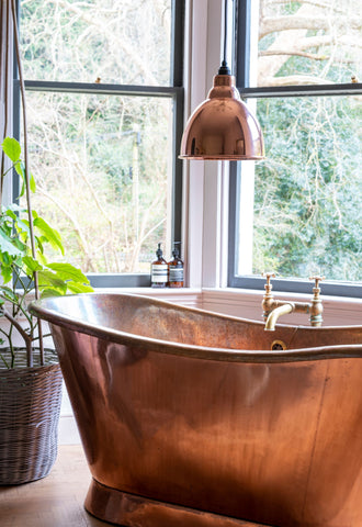 From The Anvil's Smooth Copper brindley pendant light hung over a copper bath in front of a large aluminium window, next to a small potted tree.