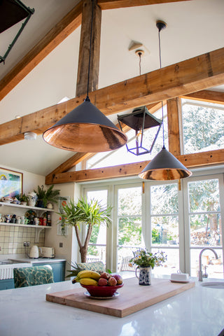 From The Anvil's Hammered Copper Hockley ceiling pendant lights hanging from wooden beams over a white kitchen island.
