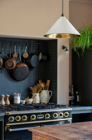A kitchen with pastel pink walls, a black tile backsplash, black oven with brass accents, pans hanging from a black rail, jugs and condiments on the side, and a Hammered Brass pendant light with a white painted exterior.