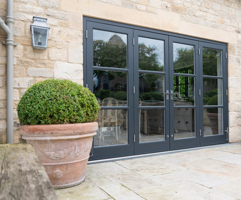 Dark grey bi-fold pation doors with From The Anvil's Pewter Avon lever door handles and butt hinges, on a stone wall with a large shrub to the side.