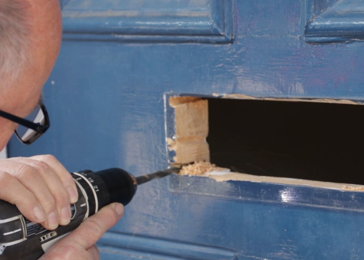 Man using an electric drill to drill a hole for a letterbox through a blue wooden door.