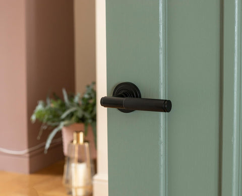 From The Anvil's Matt Black Brompton knurled lever on rose door handle on a sage green panelled door, with houseplants and a floor lamp in the background.