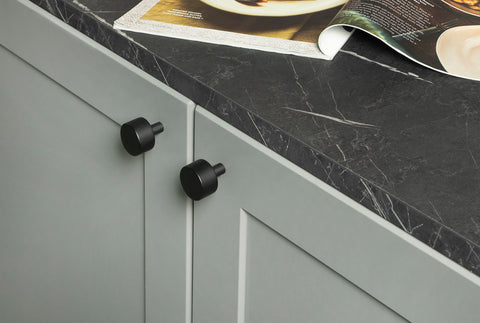 From The Anvil's Matt Black Kelso cabinet knobs on a pale green cabinet with a slate grey countertop with an open magazine on top.