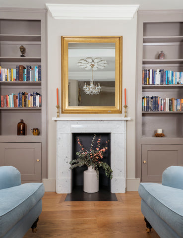 From The Anvil's Aged Brass Scully cabinet knobs on beige painted cupboards on either side of a fireplace with a gold framed mirror above it.