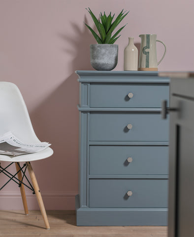 From The Anvil's Polished Nickel Kahlo cabinet knobs on a blue chest of drawers with a jug and potted plant on top, a white chair to the left, and a pink wall behind.
