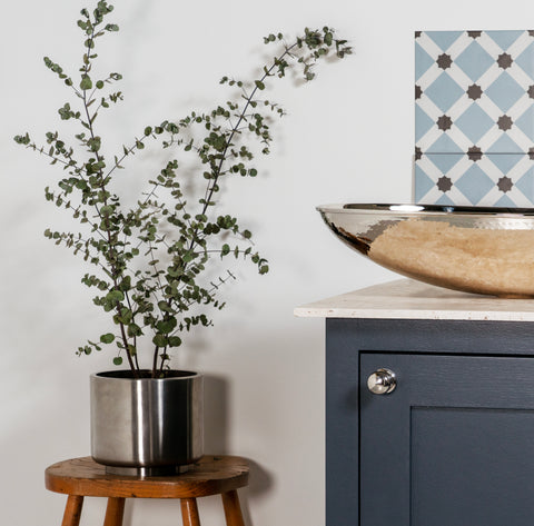 From The Anvil's Stainless Steel Newlyn plant pot with a eucalyptus plant in it, on a round wooden stool, next to a blue bathroom cabinet with a Polished Nickel sink on top.