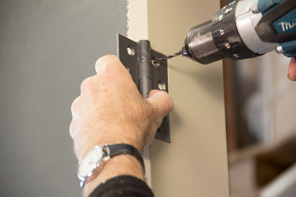 Person holding a Pewter butt hinge to a door frame and using an electric drill to drill pilot holes into the door jamb.