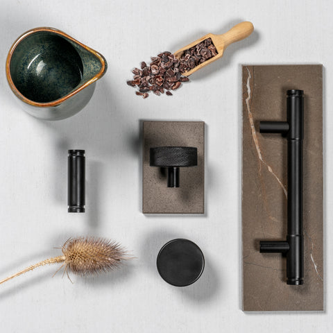 White background moodboard of From The Anvil's Aged Bronze T-bar cabinet handles, cabinet knobs, and pull handles, with brown tiles, a scoop full of coffee beans, a green jug, and a dried plant scattered around as props.