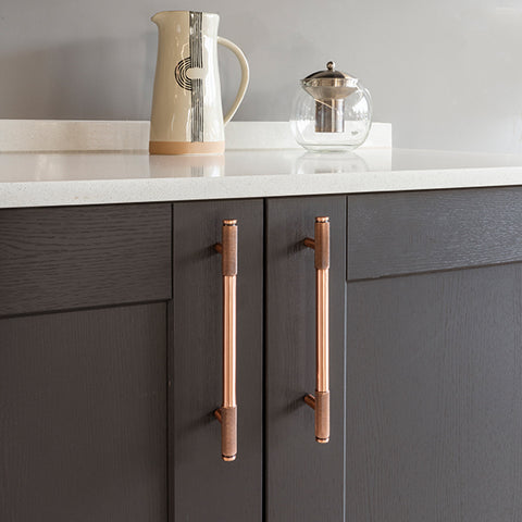 From The Anvil's Polished Bronze half Brompton knurled pull handles on a grey cabinet with a white marble counter, with a decorative jug and glass on top.