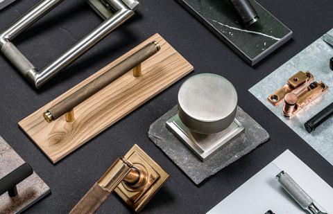 A black-background moodboard of From The Anvil's Brompton knurled pull handle, door knob, door knocker, and sash window hardware positioned on slabs of wood, slate, tile, and stone.