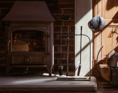 From The Anvil's Pewter Arc companion set with tongs, brush, poker, and shovel, placed in front of a stone fireplace with a black log burner (unlit).