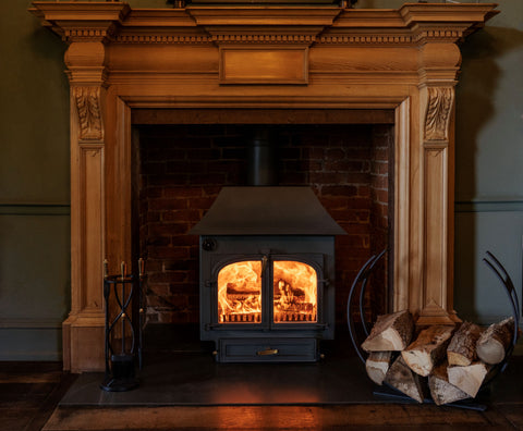 Black log burner with a blazing fire in a brick fireplace surrounded by an ornate wooden mantle, with a Black Spiral companion set and Matt Black Arc From The Anvil log holder filled with logs