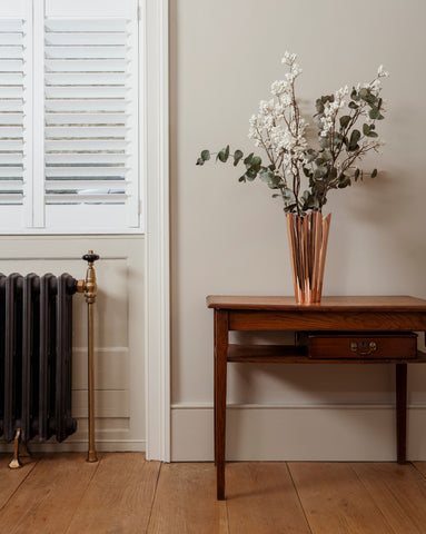 From The Anvil's Smooth Copper vase with eucalyptus and white flowers inside, on a dark wood table on a wooden floor, next to a white shuttered window with a dark grey and bronze radiator below.