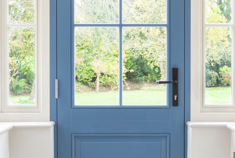 From The Anvil's Aged Bronze espag. lever lock door handle set on a blue door in a white porch with a garden visible through the windows