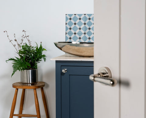 From The Anvil's Hammered Nickel sink on a white countertop on a navy blue cabinet with a Polished Nickel cabinet knob, next to a wooden stool with a potted plant on, and a painted door in the foreground with a Polished Nickel door handle.