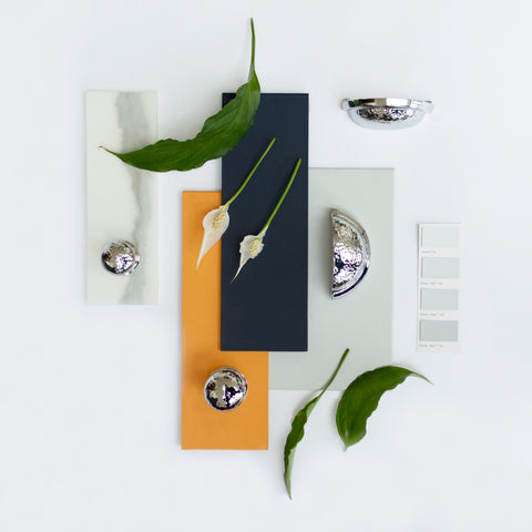 A moodboard featuring Little Greene paint samples in Marigold, Basalt, and pale grey, with From The Anvil's Polished Chrome Hammered cabinet knobs and drawer pulls placed on top. Green plant leaves have been scattered around.