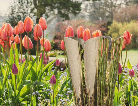From The Anvil's Polished Marine Stainless Steel Flora plant pot in a flower bed of tulips and green plants, with blurred trees in the background.