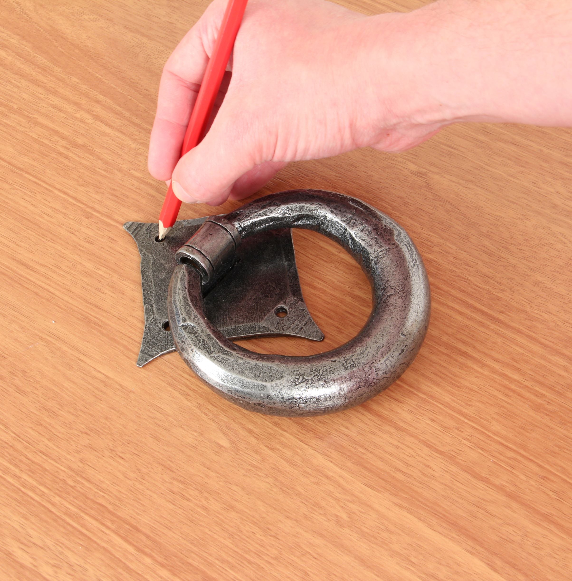Person marking the screw hole of a Pewter door knocker on a wooden door using a pencil.