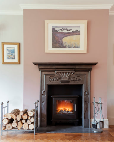 From The Anvil's Pewter log holder filled with logs, and their Pewter Spiral companion set next to an ornate fireplace with a lit log burner, and a picture frame above the mantle piece.