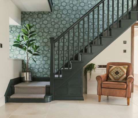 From The Anvil's Polished Stainless Steel Newlyn and Hepworth plant pots on a dark painted staircase with blue patterned wallpaper, a white stone floor, and a tan leather armchair with a tribal patterned cushion.