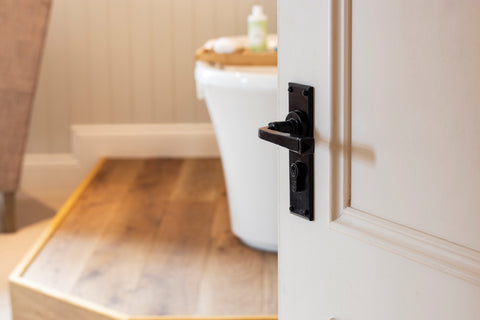 From The Anvil's Black Avon lever euro lock door handle set on a white door leading into a wood-floored bathroom