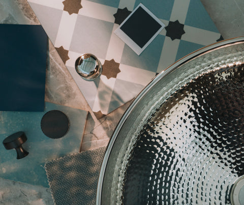 From The Anvil Hammered Nickel sink laid on a marble surface with patterned tiles, paint swatches, and cabinet knobs in a moodboard.