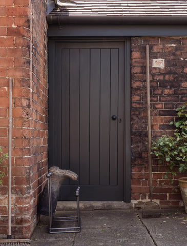 A From The Anvil Pewter boot rack with  pair of wellies on it against a red brick wall, outside a panelled wooden door with a Black From The Anvil door knob.
