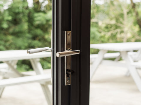 From The Anvil's Polished Stainless Steel Brompton knurled slimline lever espagnolette door handle on a grey door frame, with two benches and some bushes visible through the glass door.