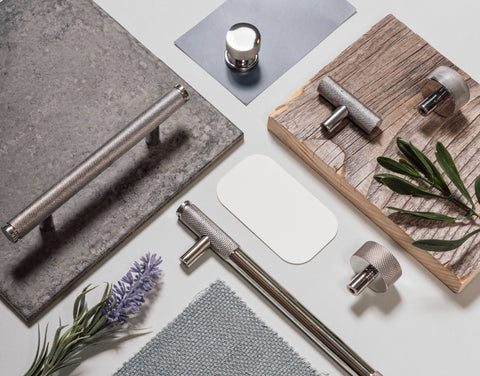 A moodboard of From The Anvil's Polished Stainless Steel hardware (including cabinet pull handles, cabinet knobs, and t-bar handles) featuring samples of grey stone, natural wood, lavender flowers, woven blue fabric, and a white rounded-square, all arranged on an off-white background.