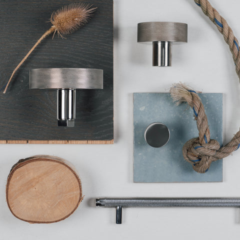 A moodboard of From The Anvil's Marine Stainless Steel hardware including their Brompton knurled door knob, cabinet knob, and matching pull handle on a white background with a blue tile, dark stained wood plank, piece of rope, slice of wood, and dried plant.