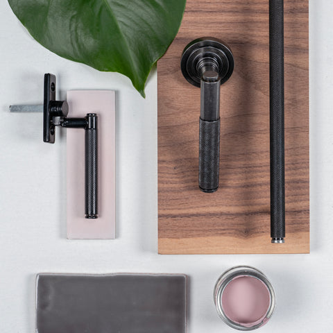 A moodboard of From The Anvil's Pewter and Black knurled hardware (lever door handle, window espag. and knurled pull handle) with painted timber, a plank of wood, a green leaf, and an open pot of pink paint.