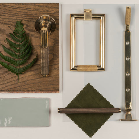 A moodboard of From The Anvil's Polished and Aged Brass Brompton knurled door knocker, lever door handle, window stay, and cabinet pull handle, laid out on a pale blue tile, dark wood plank, and green fabric square, with a green fern leaf.