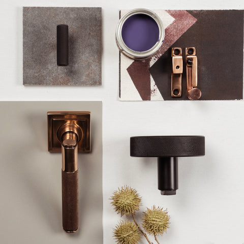 From The Anvil's Polished Bronze and Matt Black knurled Brompton cabinet handles, lever door handle, sash window hardware, and centre door knob in a neutral moodboard with an open pot of purple paint and a dried plant.