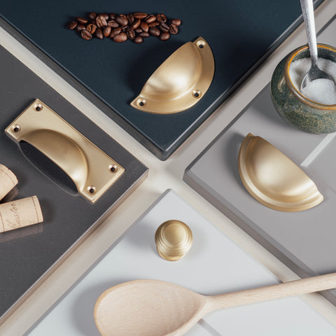 A kitchen-themed moodboard of From The Anvil's Satin Brass hardware, featuring 3 drawer pulls and one cabinet knob. Each product is on a different coloured cabinet next to a matching kitchen prop (wine corks, wooden spoon, jar of sugar, coffee beans).