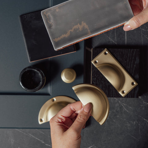 A moodboard of From The Anvil's Satin Brass drawer pulls and cabinet knobs contrasting with black tiles, dark wood, grey tiles, an open tester pot of black paint, and a navy blue kitchen cabinet.