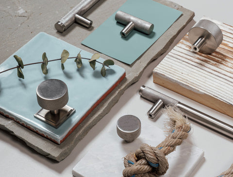 A moodboard of From The Anvil's Satin Marine Stainless Steel knurled Brompton hardware including knurled cabinet knobs, pull handles, and T-bars. The hardware is aesthetically placed on blue tiles, pieces of slate, and unfinished wood. Eucalyptus leaves and jute rope have been added as props.