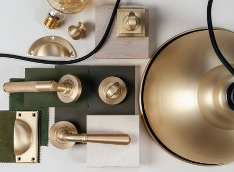 From The Anvil's Satin Brass hardware & lighting moodboard with green paint & fabric samples, wooden & marble tiles, and Satin Brass pendants, lever door handles, thumbturns, a drawer pull, and a lightbulb.