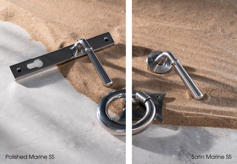 A white marble countertop with sand laid out in a wave pattern. On the sand is From The Anvil's Polished SS Newbury slimline lever lock handle, Satin SS Newbury lever on rose door handle, and a Ring door knocker. The image is split down the middle (vertically) - on the left side is the Polished SS hardware, and on the right side is the Satin SS hardware. The Ring door knocker is crossed by this line, so it is half Polished SS and half Satin SS.