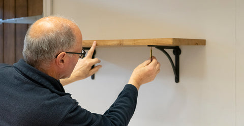 Man using a pencil to mark the screw holes on the underside of a wooden shelf.