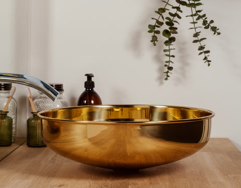 From The Anvil's Smooth Brass Round sink on a wooden countertop, chrome tap, and assorted glass jars in the back and a green plant trailing down above it.
