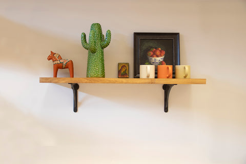 A wooden shelf hung using From The Anvil's Beeswax Curved shelf brackets with assorted ornaments and ceramic mugs on top.