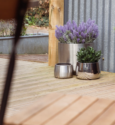 From The Anvil's Satin and Polished Marine Stainless Steel Newlyn and Hepworth plant pots with lavender and green plants on an outdoor decking area.