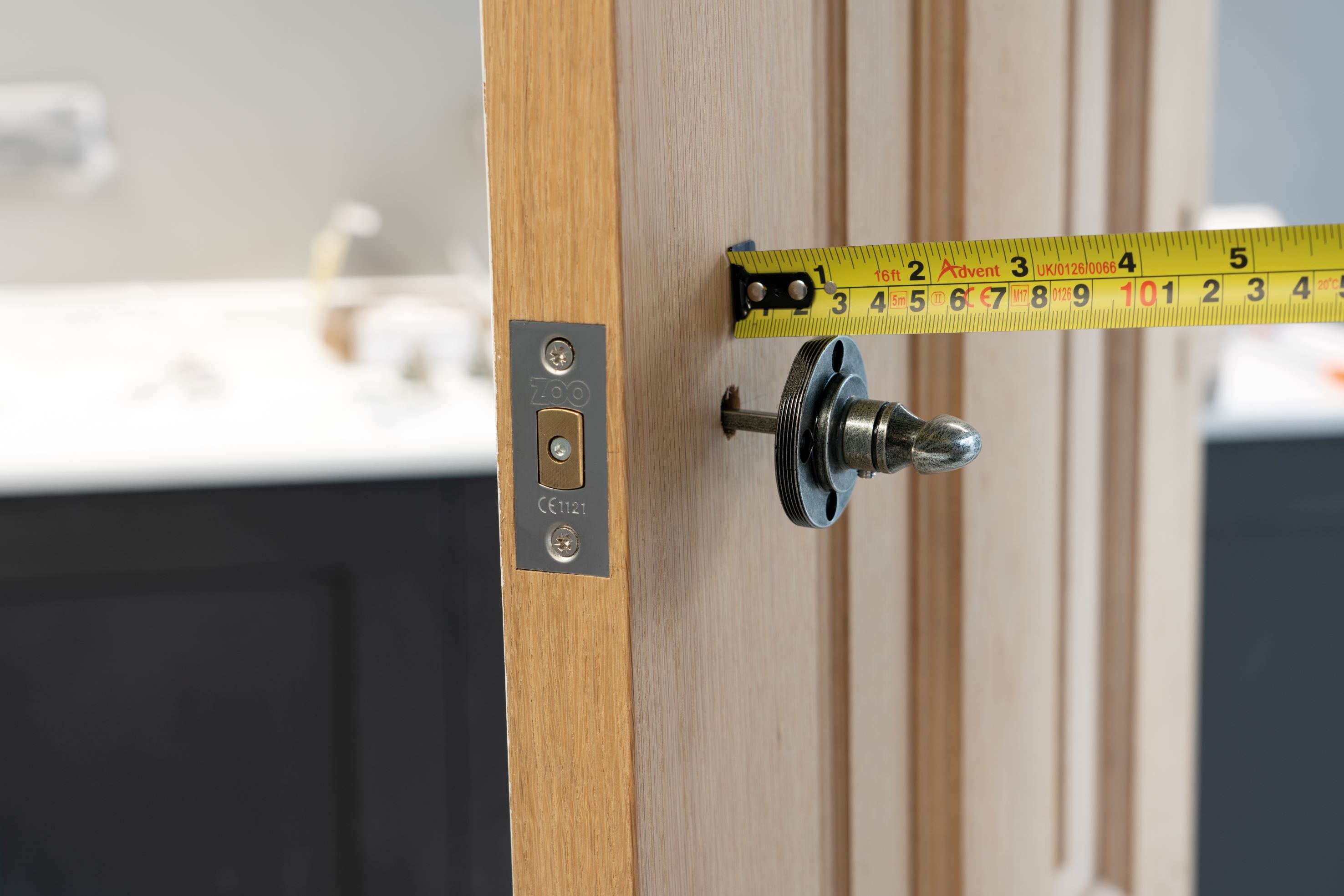 A Pewter thumbturn being attached to a spindle passing through a wooden door, with a tape measure measuring the gap between the door and the thumbturn