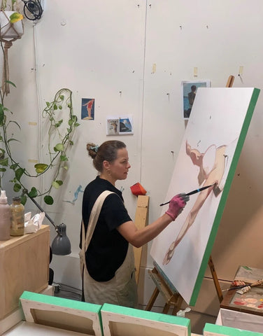 A caucasian woman in her 40s with brown hair in a bun, wearing a painting smock and painting at an easel in an art studio