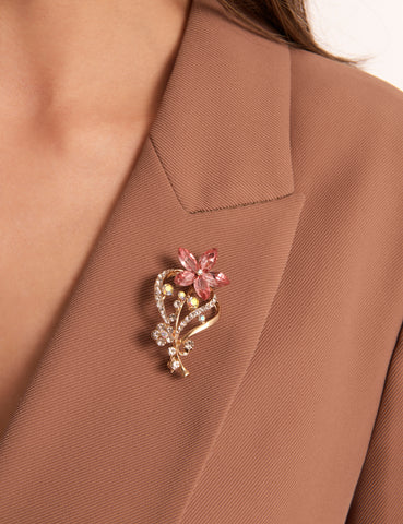 Close-up of a woman wearing a tan blazer with the Lilith brooch. It features a flower design with pink stone petals, gold-coloured plating and sparkling stone details.