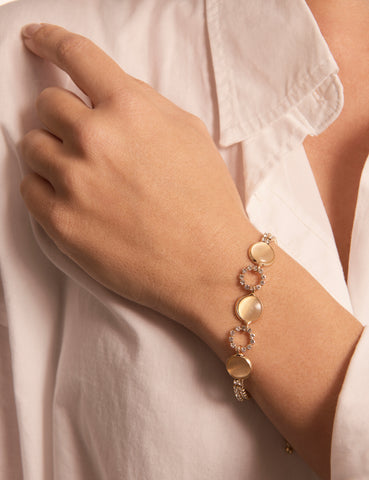 The Saturn Bracelet on a woman's wrist. It is a gold coloured braclet, with round natural-toned stones, circular links and fine sparkling stones. The woman's hand lays across her chest. She is wearing a white shirt.