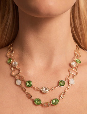 Close-up of a woman's neck. She is wearing the Double Stone Shape Necklace with faceted green and white stones and gold-coloured plating. She has pale skin and blonde hair.